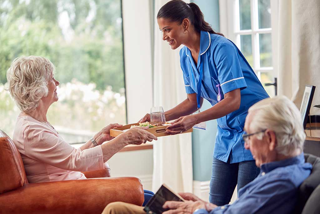 Female Care Worker In Uniform Bringing Meal On Tray To Senior Woman Sitting In Lounge At Home